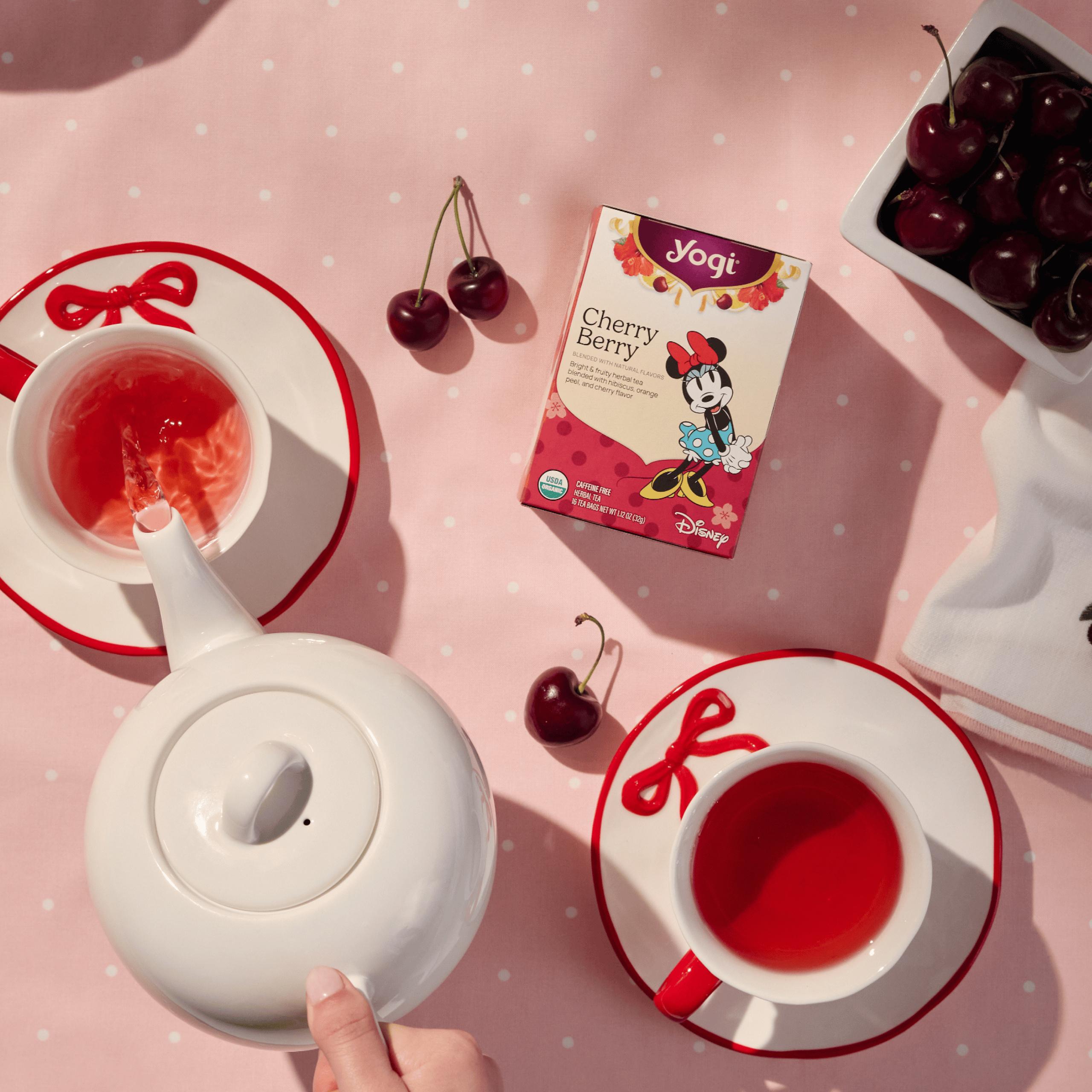 A person's hand gently pouring vibrant red Yogi Cherry Berry tea from a white teapot into a matching teacup