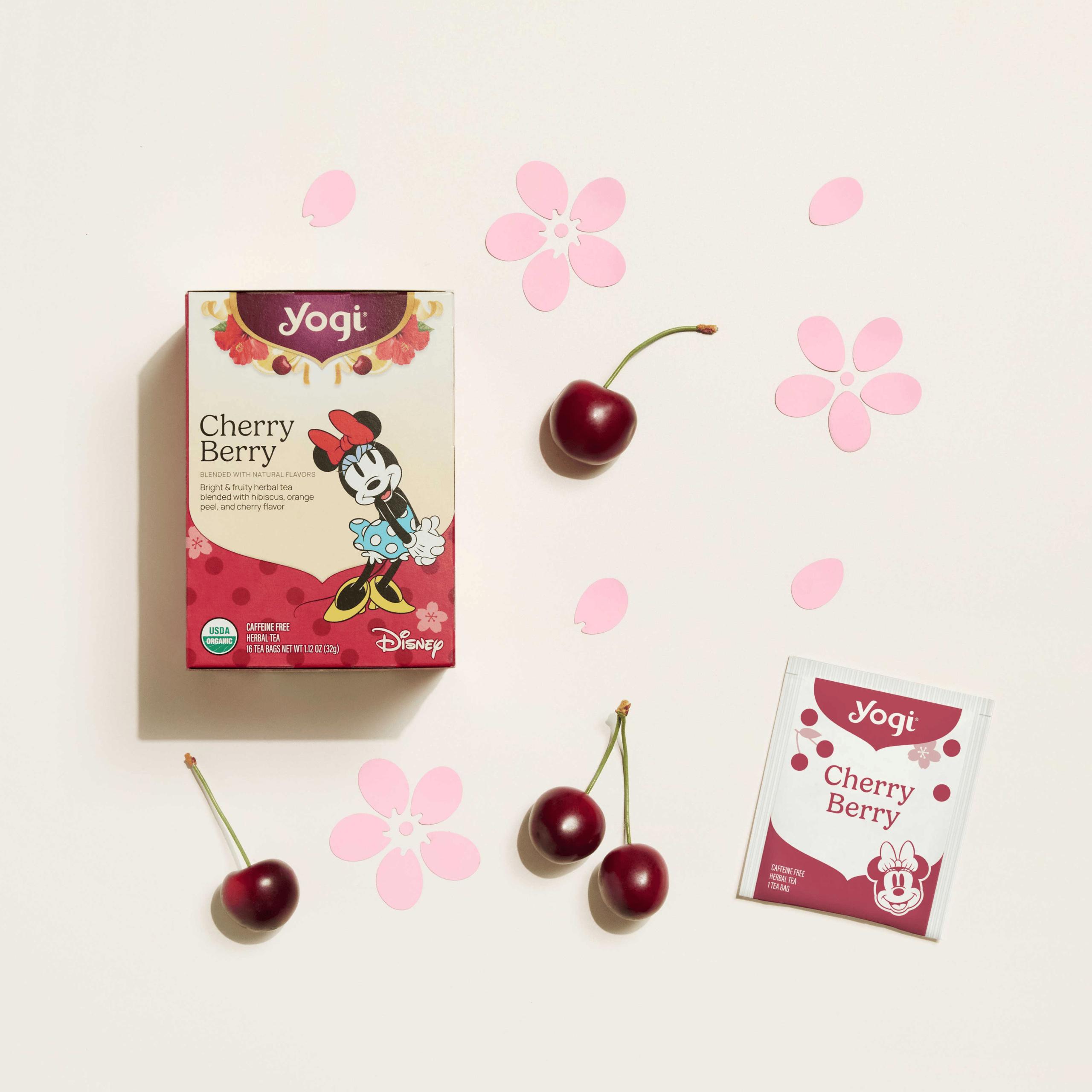 A top-down view of Yogi Cherry Berry tea packaging and an individual tea envelope on a soft cream background with fresh cherries and pink flower cutouts