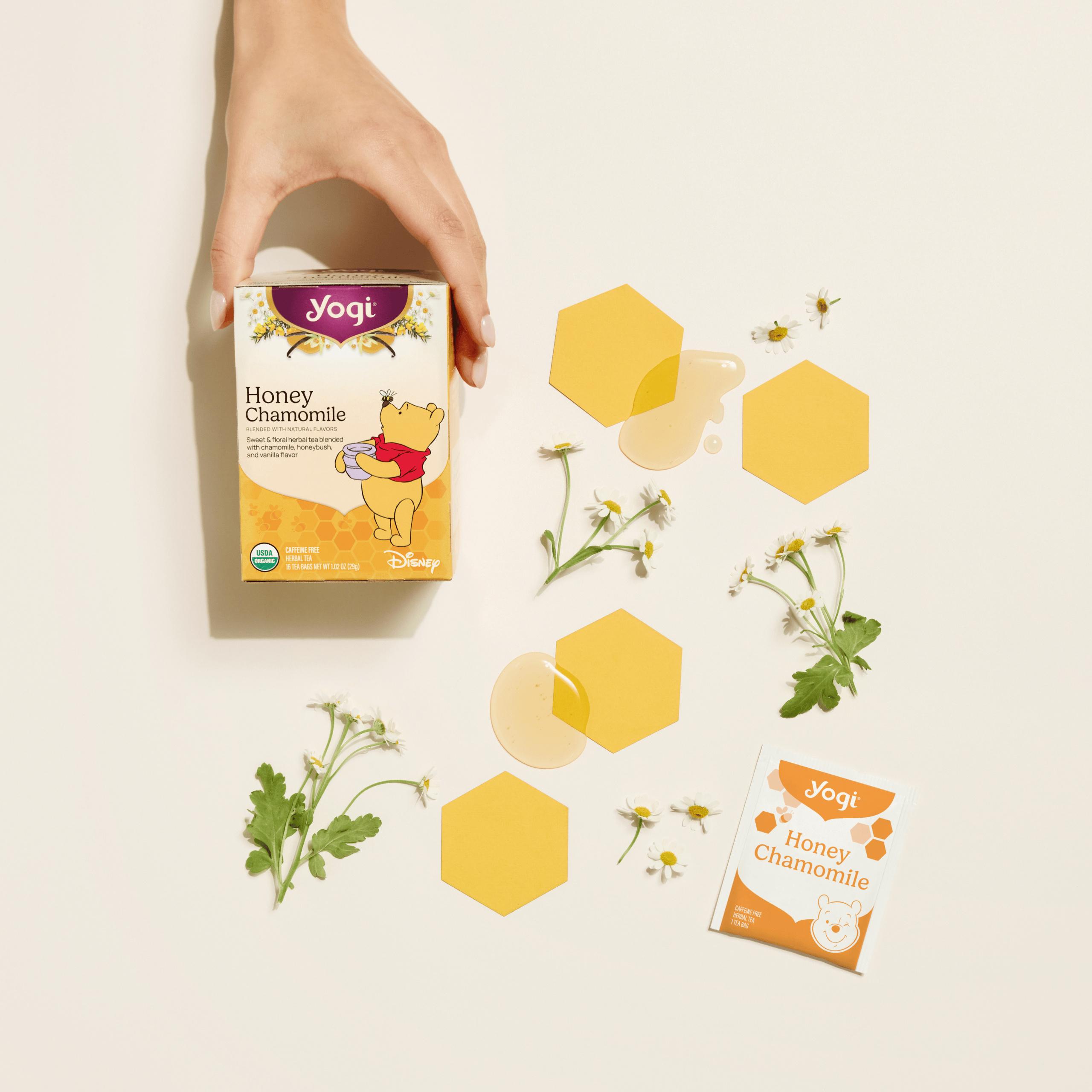 A top-down view of a hand placing a box of Yogi Honey Chamomile tea onto a cream-colored surface, decorated with individual tea envelopes and fresh chamomile sprigs