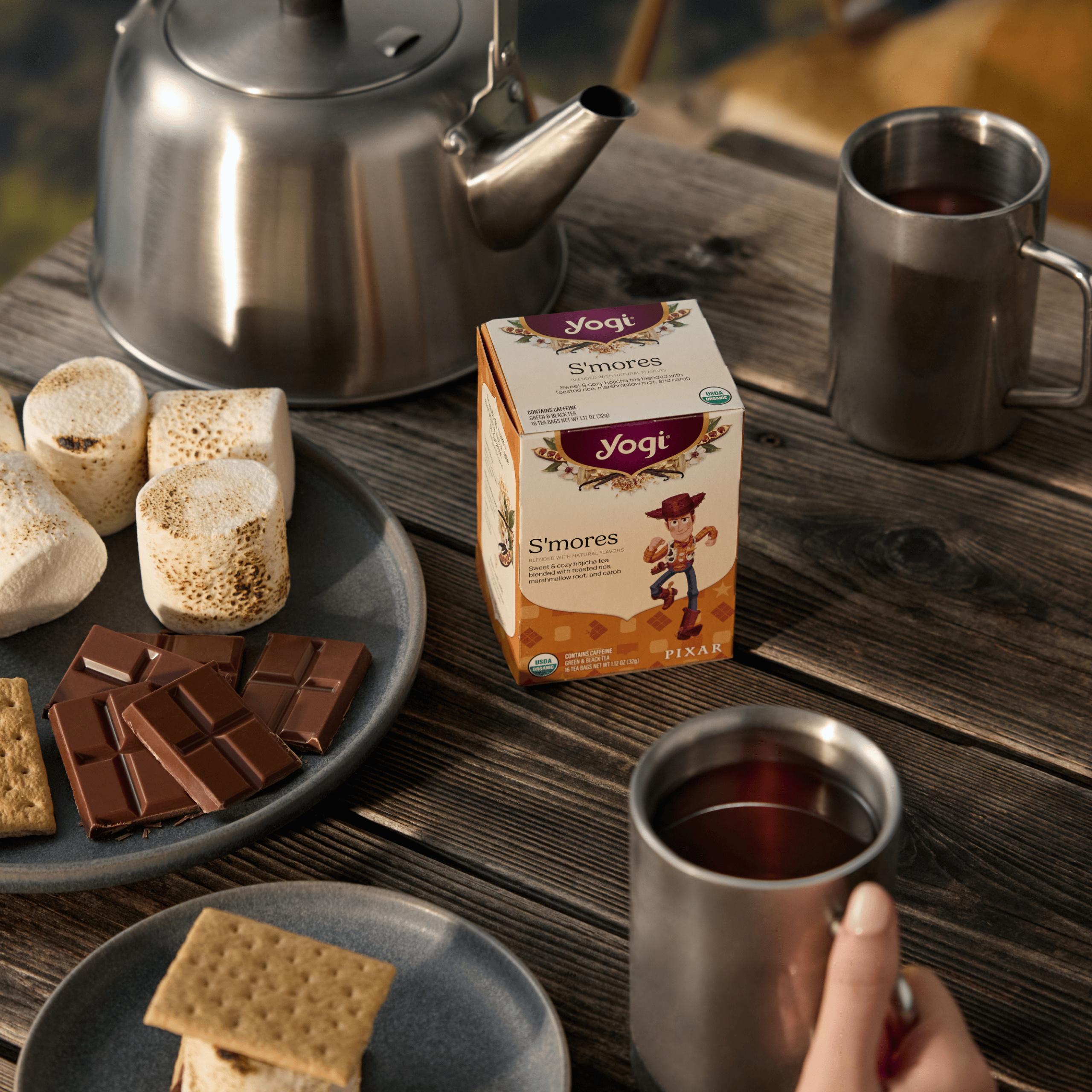 A cozy outdoor scene on a wooden table featuring Yogi S’mores tea, with a plate of s'more ingredients, a silver kettle, and a hand holding a steel mug of tea