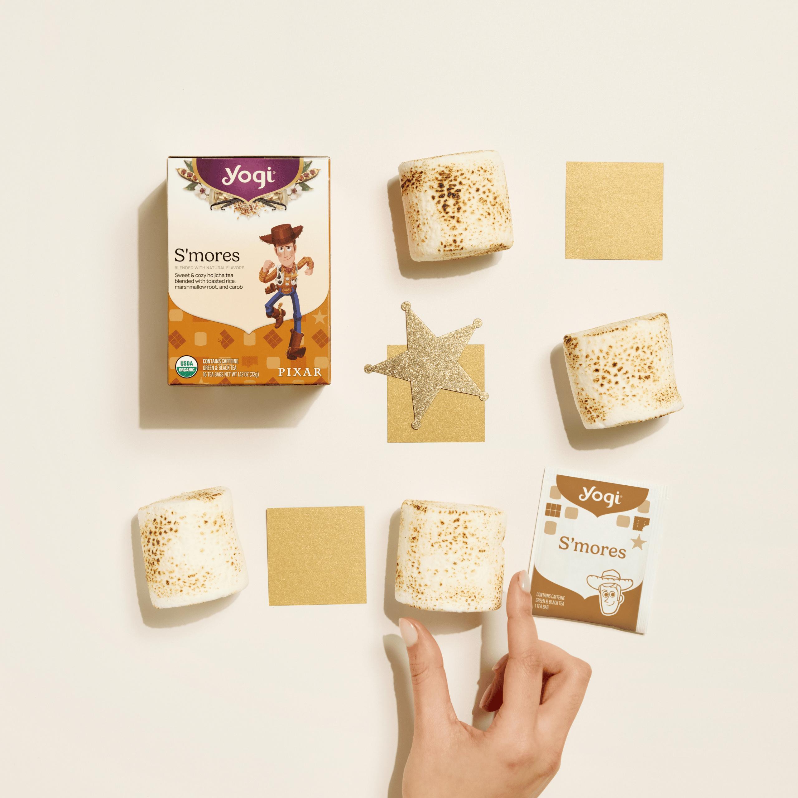A top-down view of Yogi S’mores tea packaging and a single tea envelope on a warm cream background, arranged with large marshmallows and a hand reaching in
