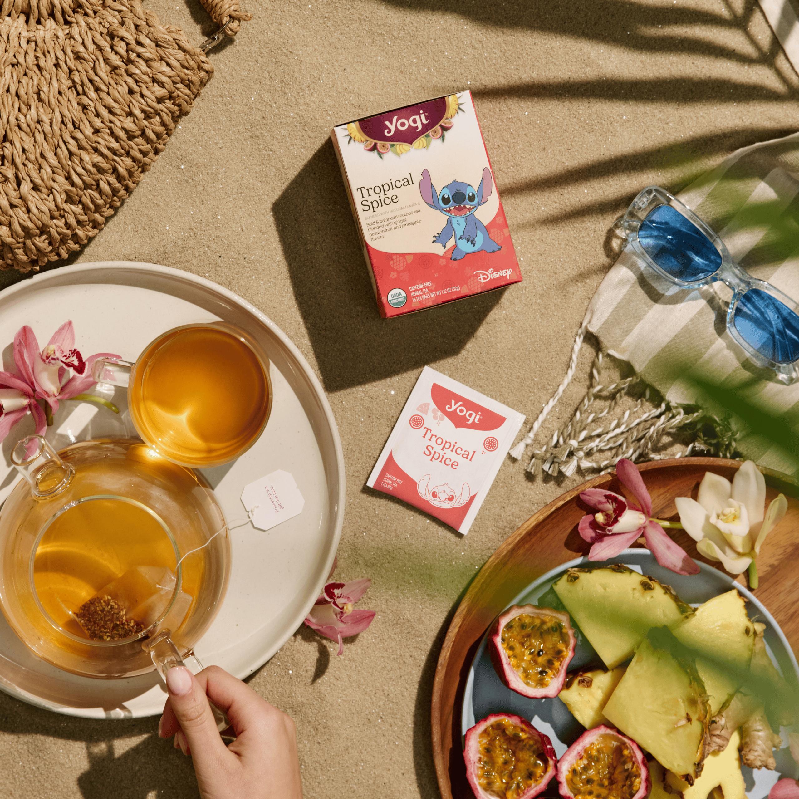 A sunny beach picnic scene on sand with a box of Yogi Tropical Spice tea, a hand holds a clear glass teapot next to a cup of golden tea, fresh passion fruit and pineapple