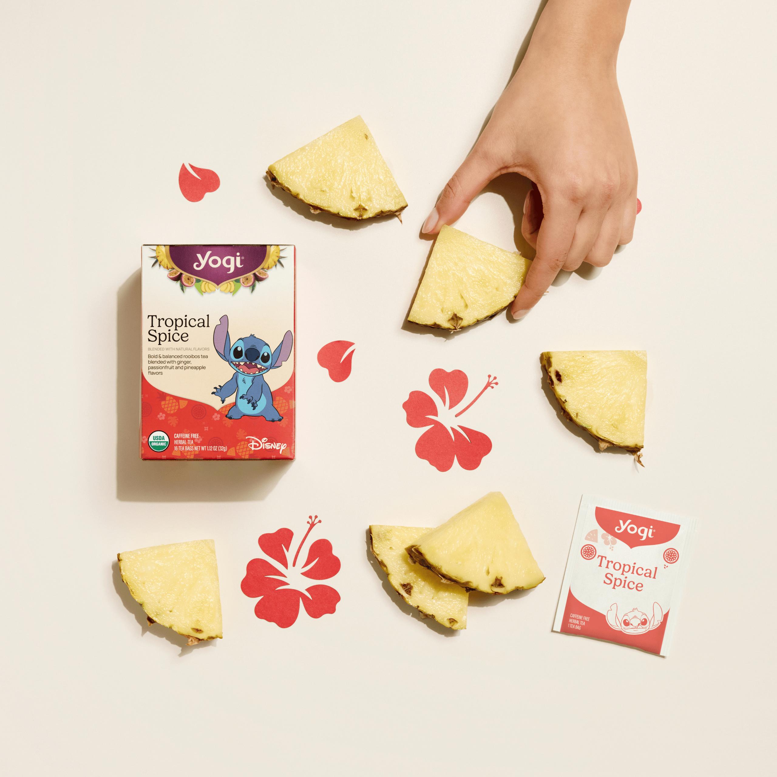 A top-down view of Yogi Tropical Spice tea box and a single tea envelope are set against a cream background, arranged with fresh pineapple wedges and vibrant red hibiscus flower paper cutouts