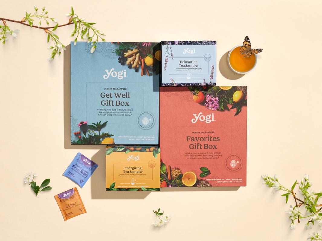 High-angle lifestyle shot of Yogi Tea Spring Promotion gift sets. Featured products include the Get Well Gift Box (nine tea blends for immune support) and the Favorites Gift Box (nine beloved tea blends), each containing 45 tea bags. Smaller samplers for Relaxation and Energizing teas are pictured alongside individual Honey Lavender Stress Relief and Ginger tea envelopes, surrounded by floral branches and a butterfly.