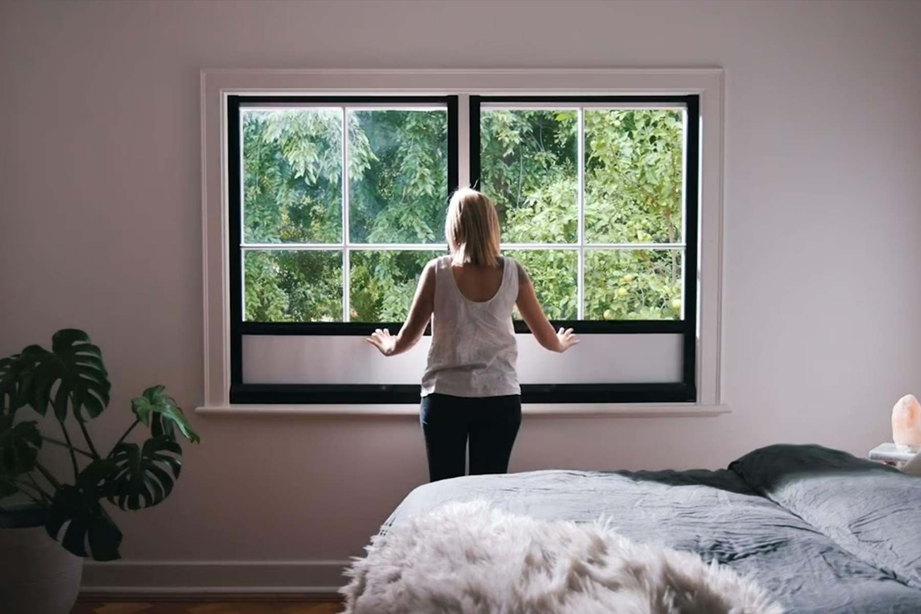 Woman standing at bedroom window with UBlockout top down bottom up shades raised from bottom for privacy while allowing natural light from top