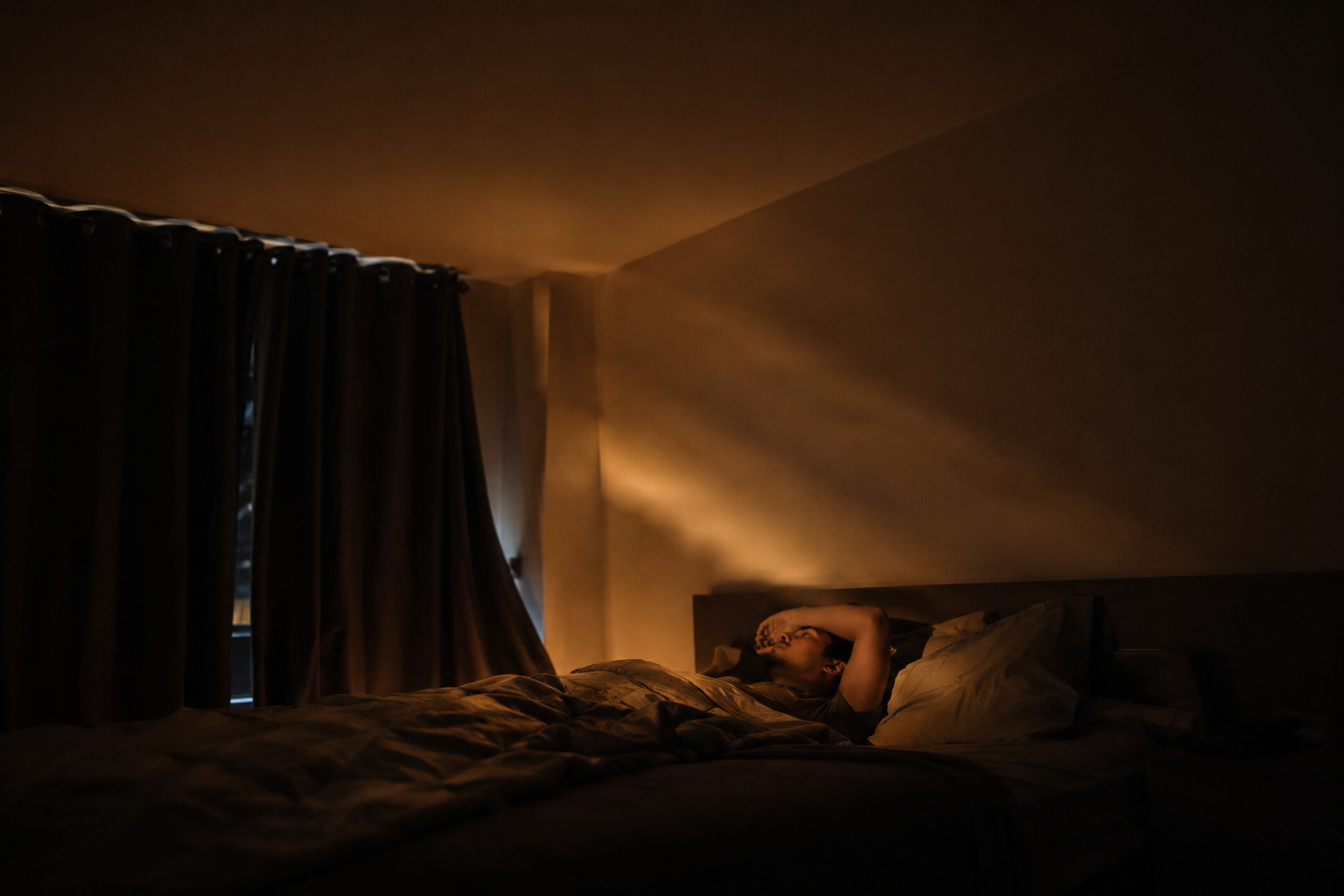 Dark urban bedroom before dawn with a restless person lying awake as passing headlights cast a warm amber sweep across the ceiling and wall. Heavy curtains leak subtle light at the edges, while the rest of the room stays deep, cinematic, and shadowed.