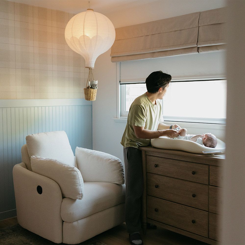  A dad changing his newborn's diaper beside a window with a UBlockout shade in a warm nursery.