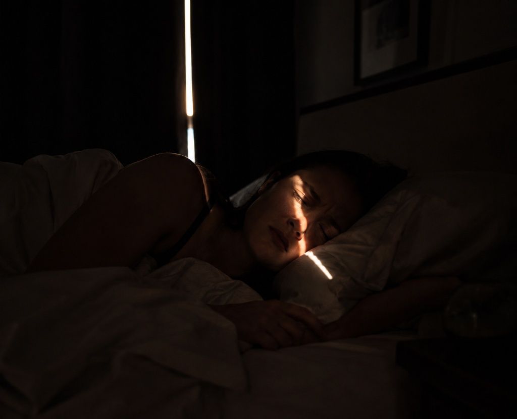 Thin line of bright morning light cutting through a gap in window coverings onto a sleeping person's face