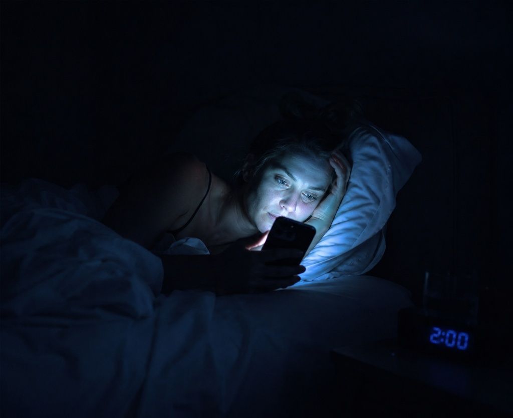 Person in bed checking their phone at 2am in a dark bedroom, face lit by screen glow