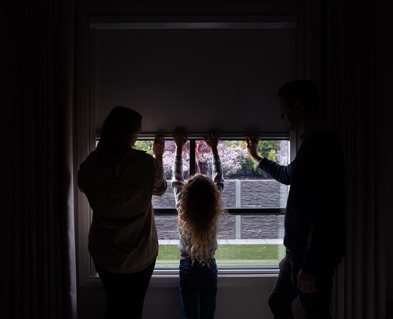 Family silhouetted against window with room darkening shade partially raised showing how figures are visible through shaded glass