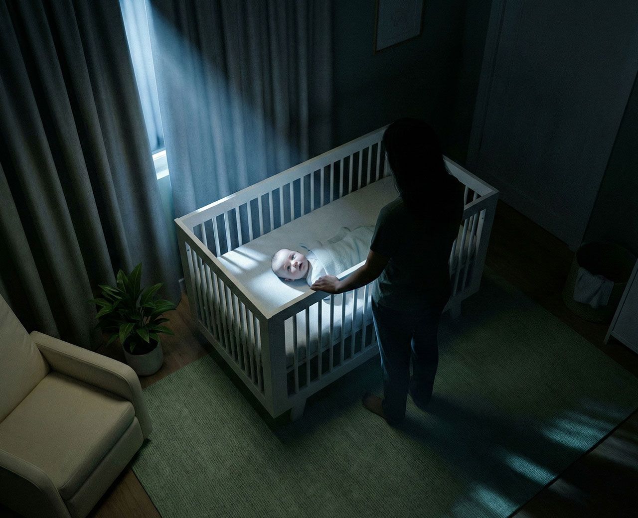 Parent standing over baby in crib at night with light leaking through curtains
