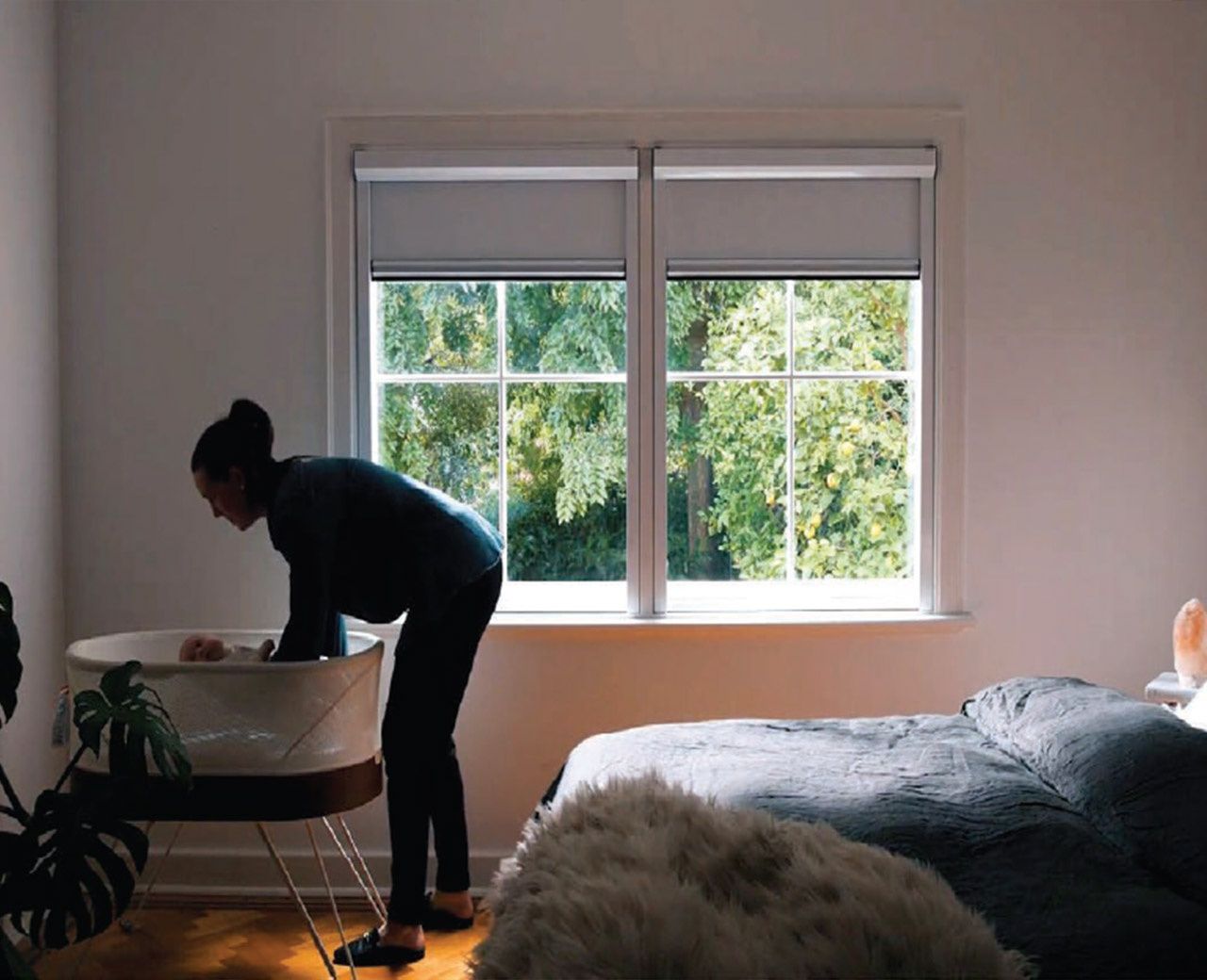 Mother tending to baby in bassinet next to window with partially raised roller shades letting in daylight