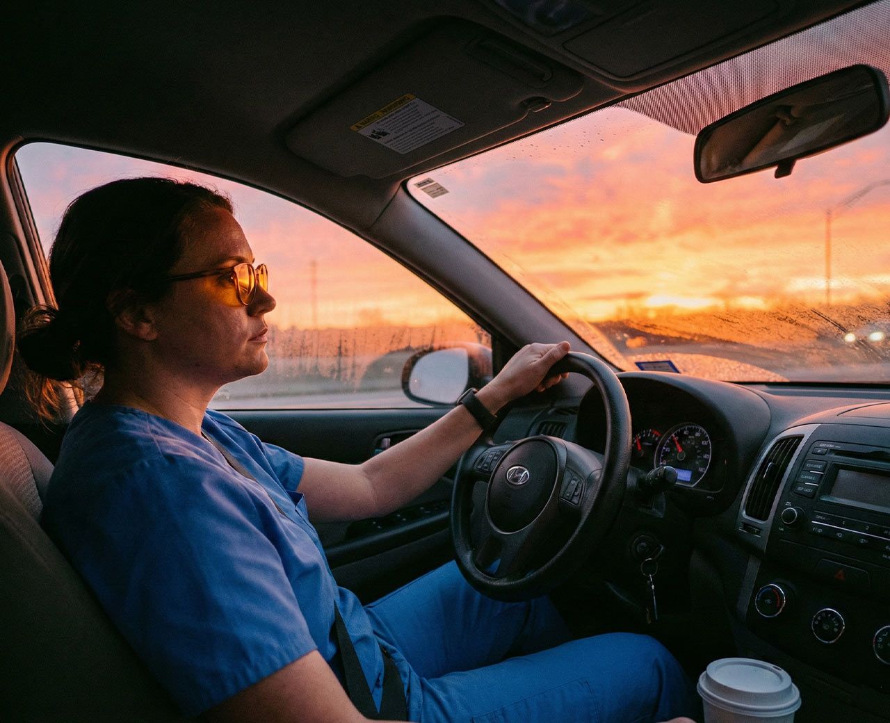 Night shift nurse in scrubs wearing blue-blocking glasses while driving home at sunrise