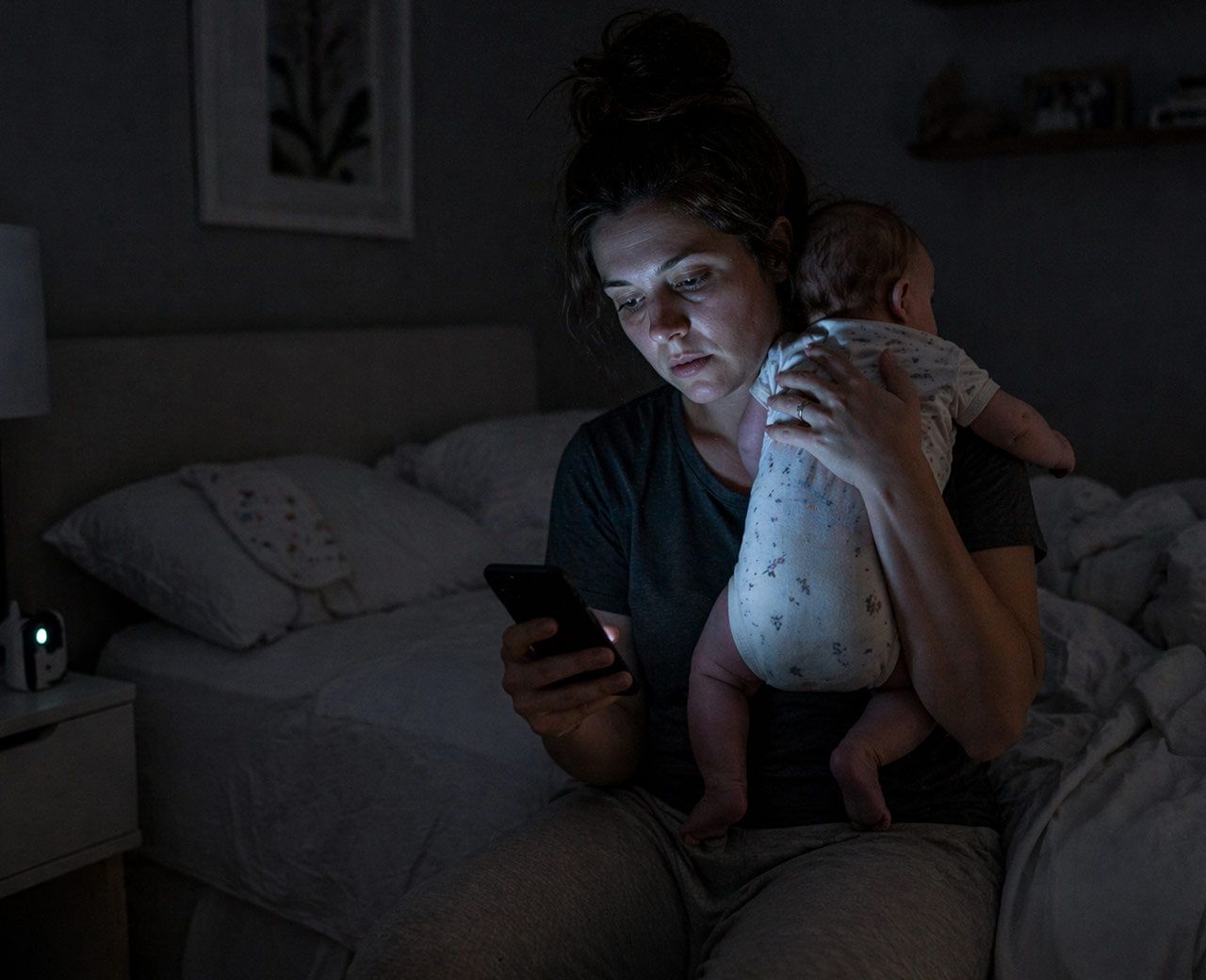 Tired mother holding awake baby at night while looking at phone in dim bedroom