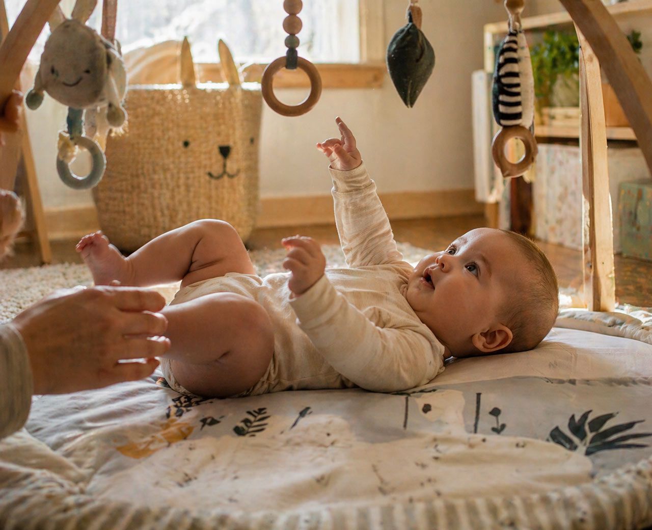 Alert baby lying on play mat looking at hanging toys with parent’s hand nearby in daylight