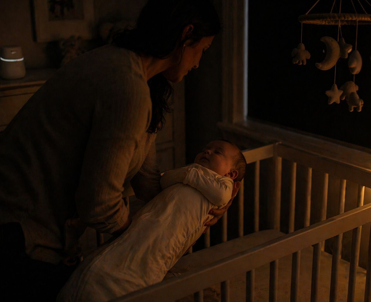 Parent placing sleepy baby into crib in dark nursery with soft amber night light