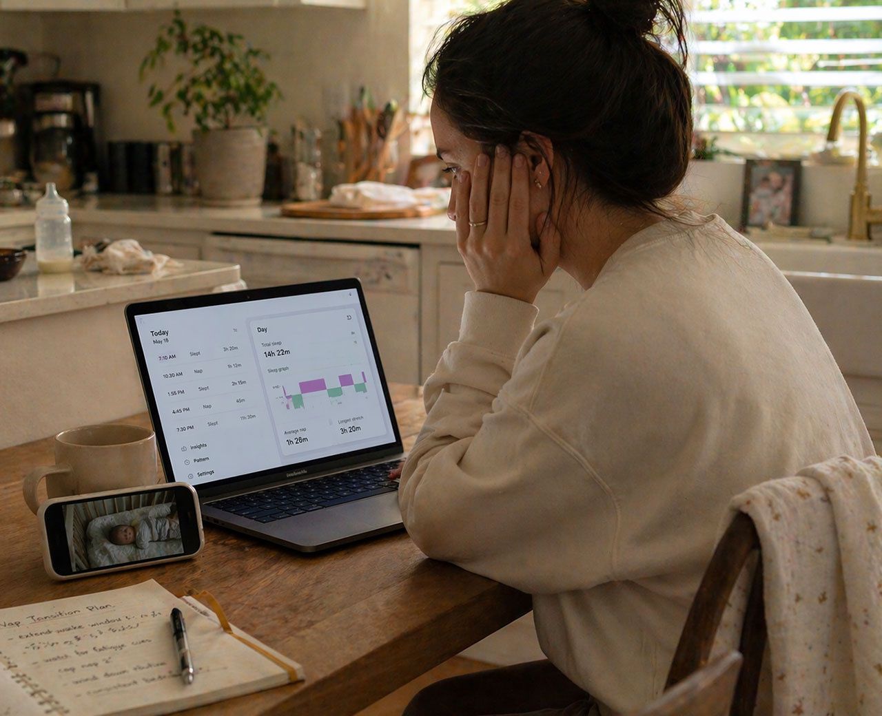 Tired parent using laptop sleep tracker at kitchen table with baby monitor and coffee nearby