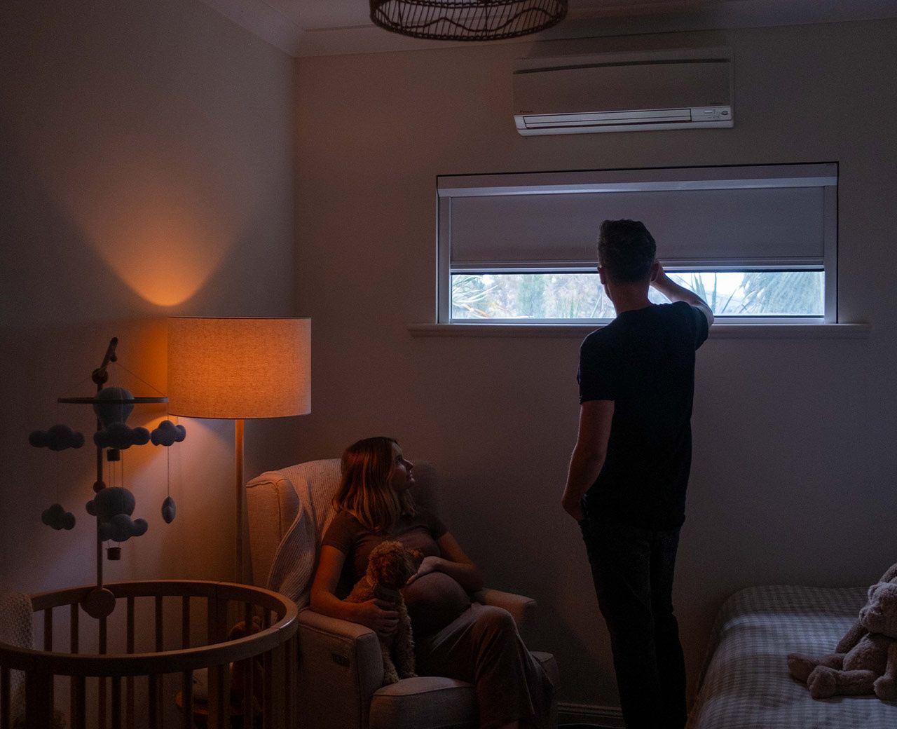 Man lowering uBlockout blackout shade in dim nursery while woman sits beside crib