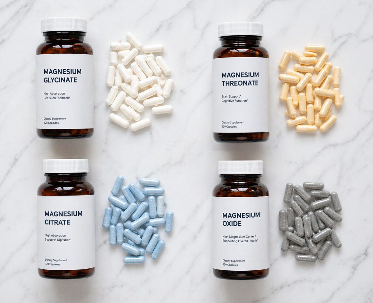 Flat-lay of four magnesium supplement types—glycinate, threonate, citrate and oxide—with different colored capsules arranged on white marble