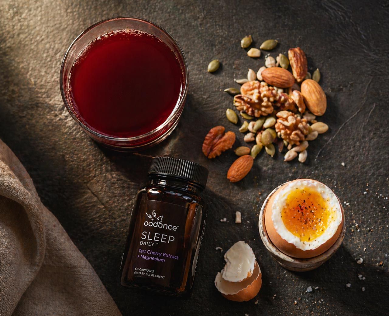 Flat-lay of tart cherry juice, supplement bottle, nuts and soft boiled egg on dark surface