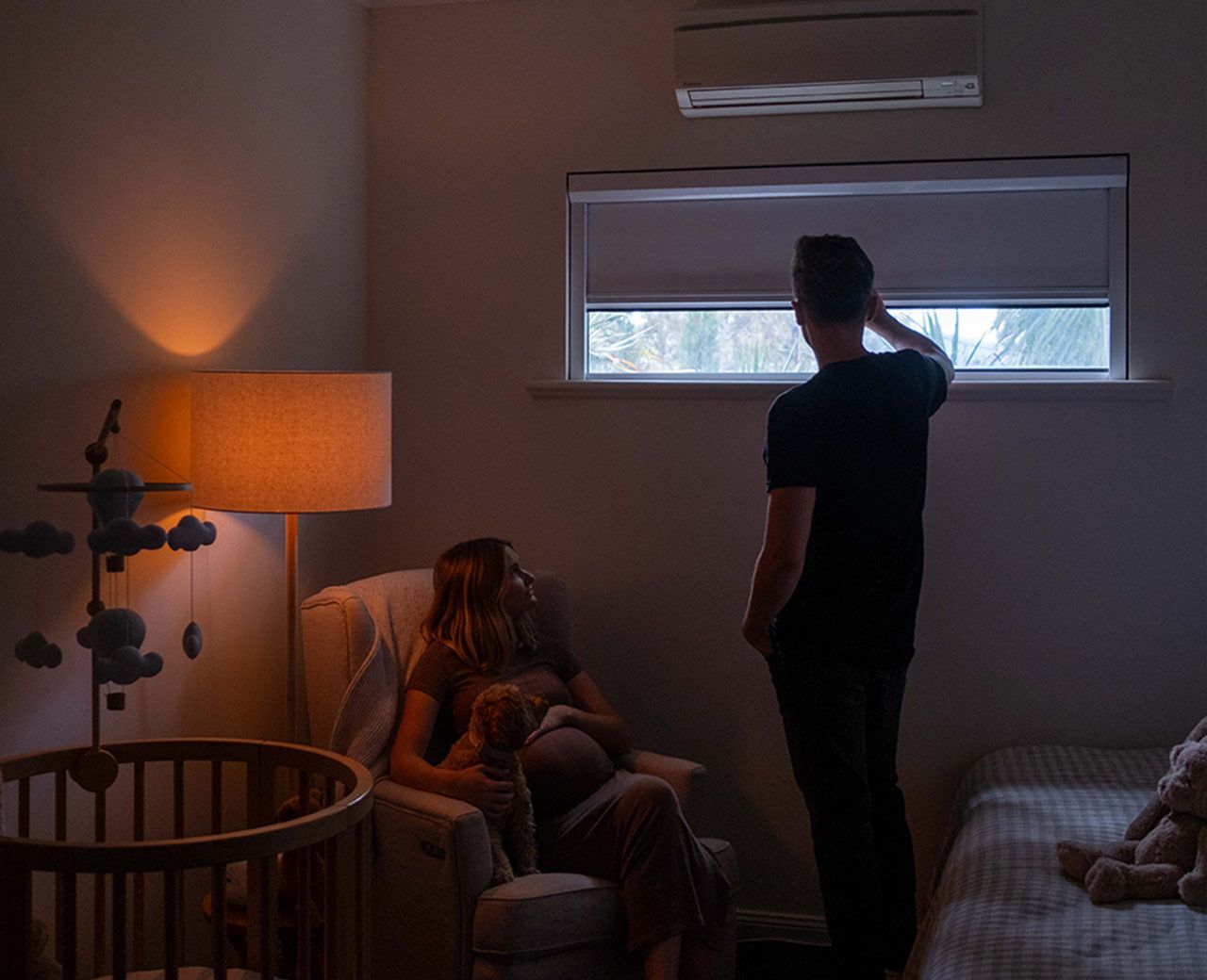 Man adjusting uBlockout shade in dim nursery while pregnant woman sits nearby in warm low light
