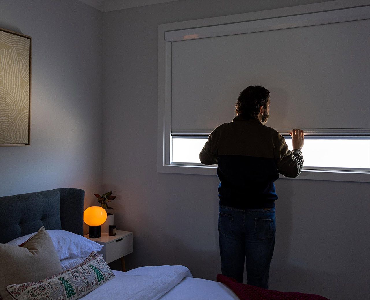 A man adjusting a UBlockout blackout shade in a dimly lit bedroom with a warm bedside lamp.