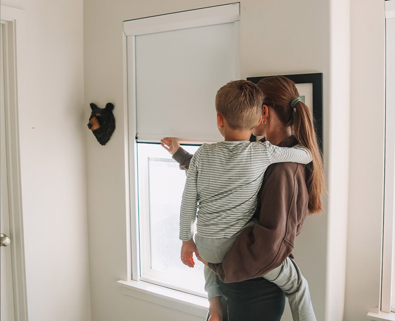 A mother holding her toddler from behind as they look at a UBlockout blackout shade on a bedroom window.