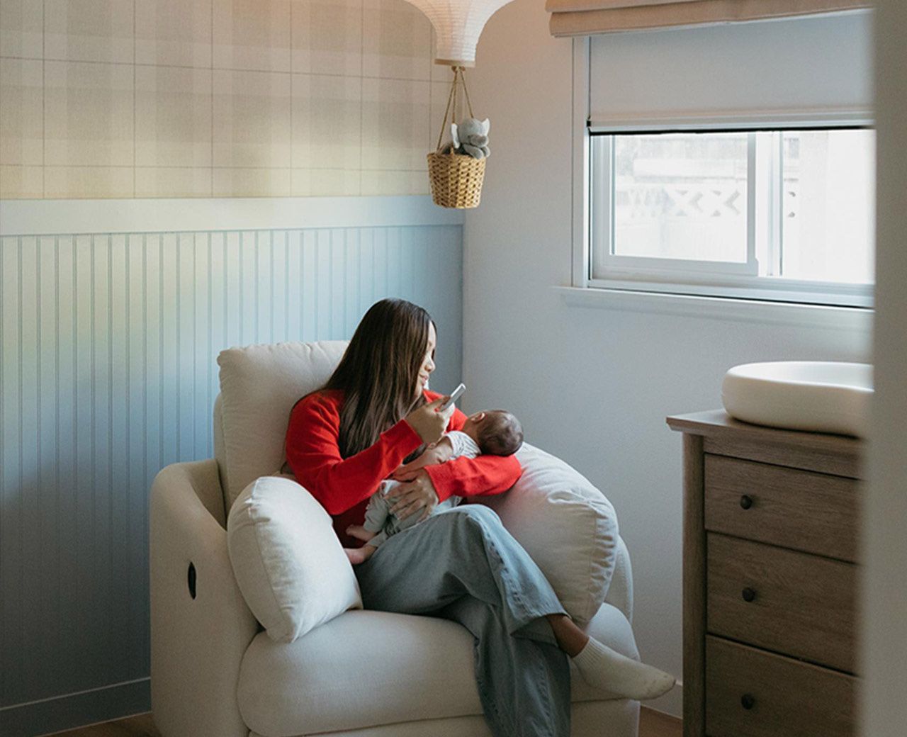 A mother sitting in a nursery recliner holding her newborn with a UBlockout shade partially covering the window behind her.