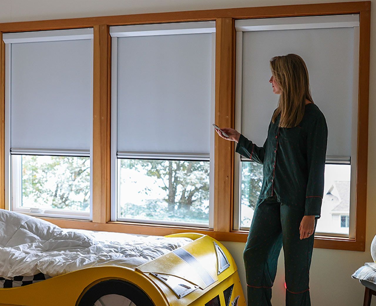 A woman in pajamas using a remote to adjust UBlockout blackout shades across three windows in a child's bedroom.
