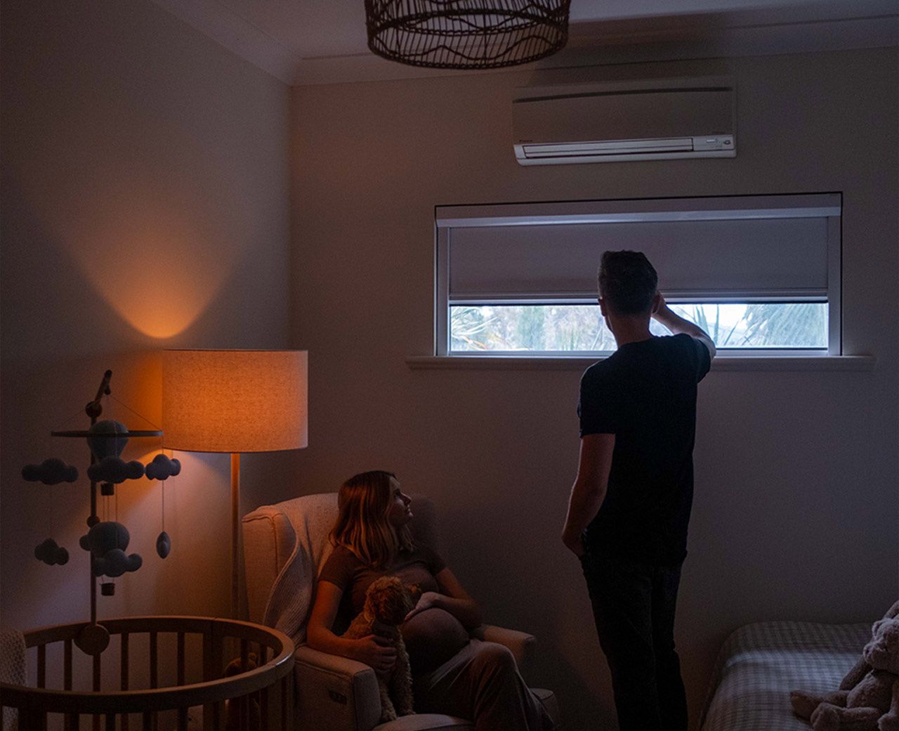 A couple in a dimly lit nursery with a man adjusting a blackout shade on the window while a woman sits in a chair holding a toy beside a cot.