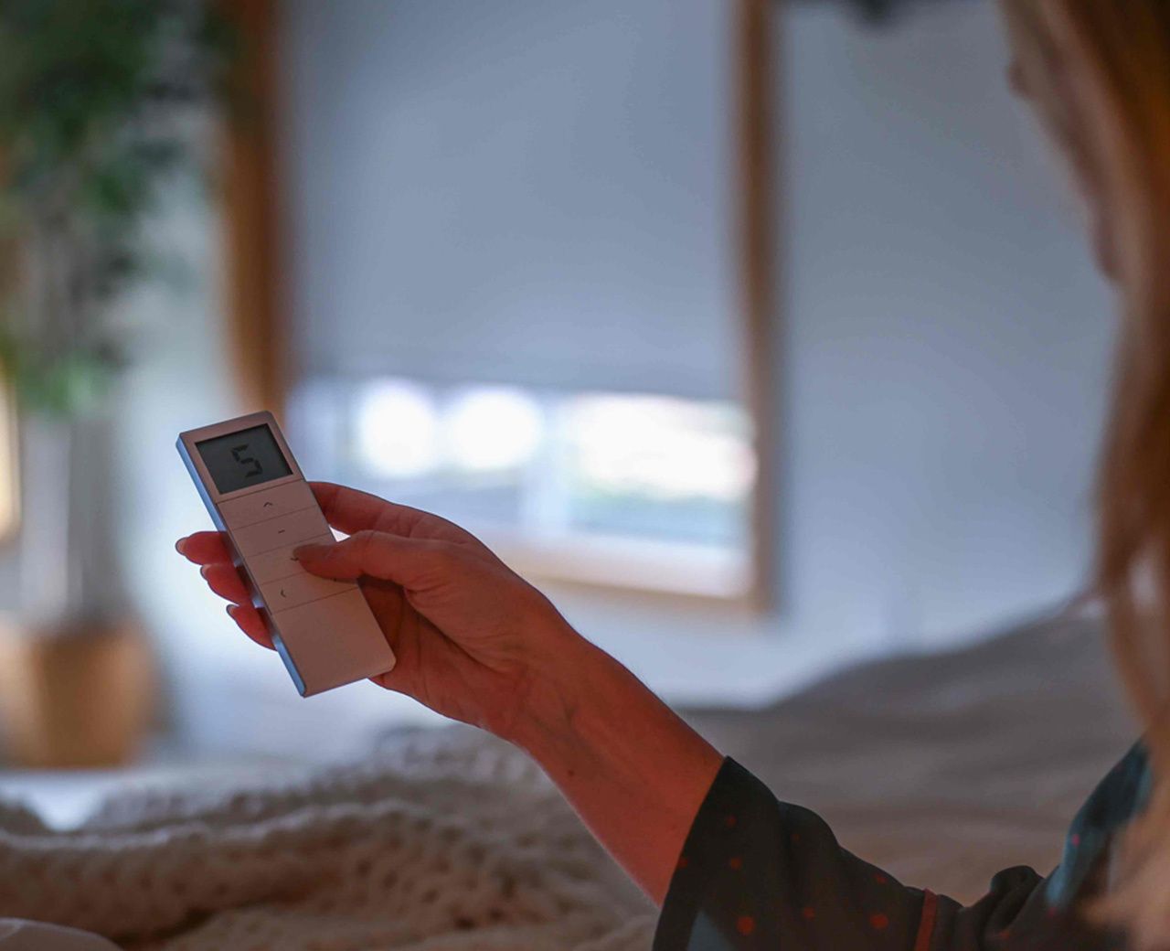 A close up of a hand holding a UBlockout motorized shade remote control with a blackout shade and timber-framed window visible in the background.