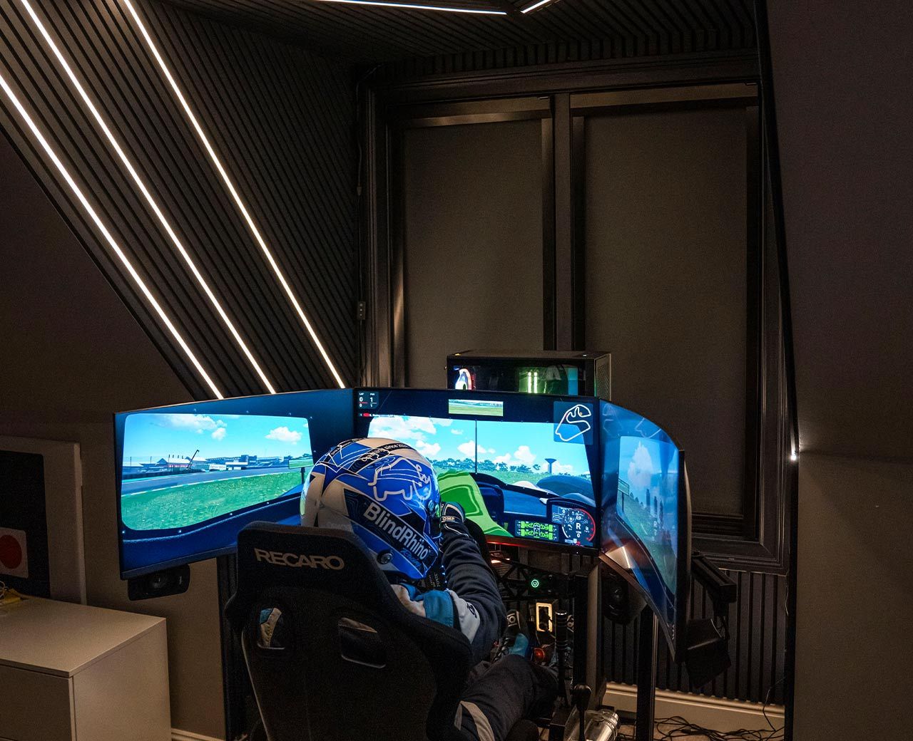  A racing simulator setup with triple screens and a helmeted driver in a darkened media room with UBlockout blackout shades installed on the window behind the rig.