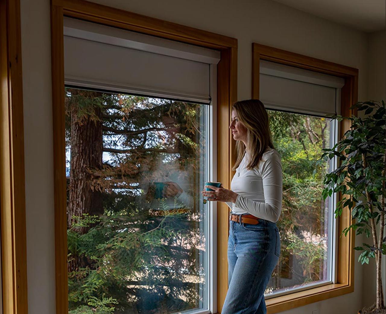 A woman holding a coffee cup looking out through two timber-framed windows with UBlockout blackout shades partially raised showing a tree-filled view outside.
