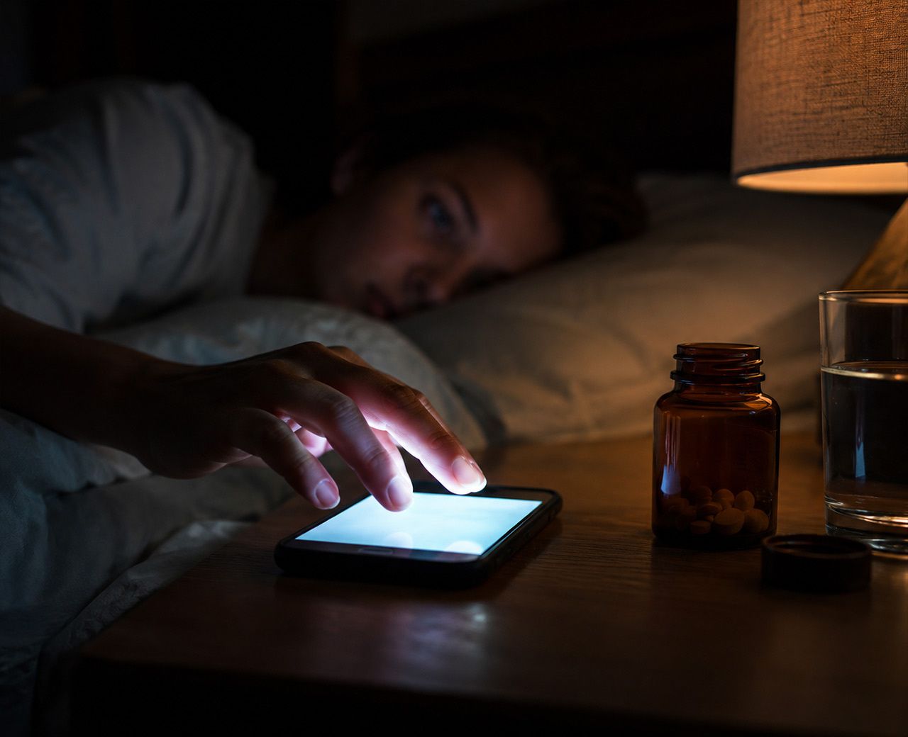 A hand reaching for a bright phone screen in a dark bedroom with a supplement bottle on the nightstand.