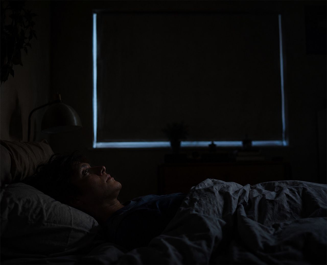 Person lying awake in a dark bedroom with faint light leaking around the edges of the window curtains