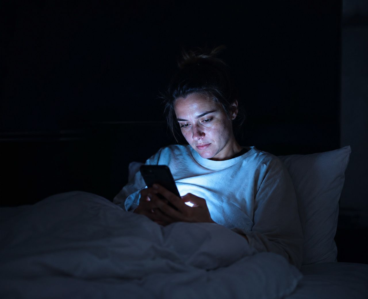 Person sitting up in bed scrolling their phone at night, face illuminated by blue screen light in an otherwise dark bedroom