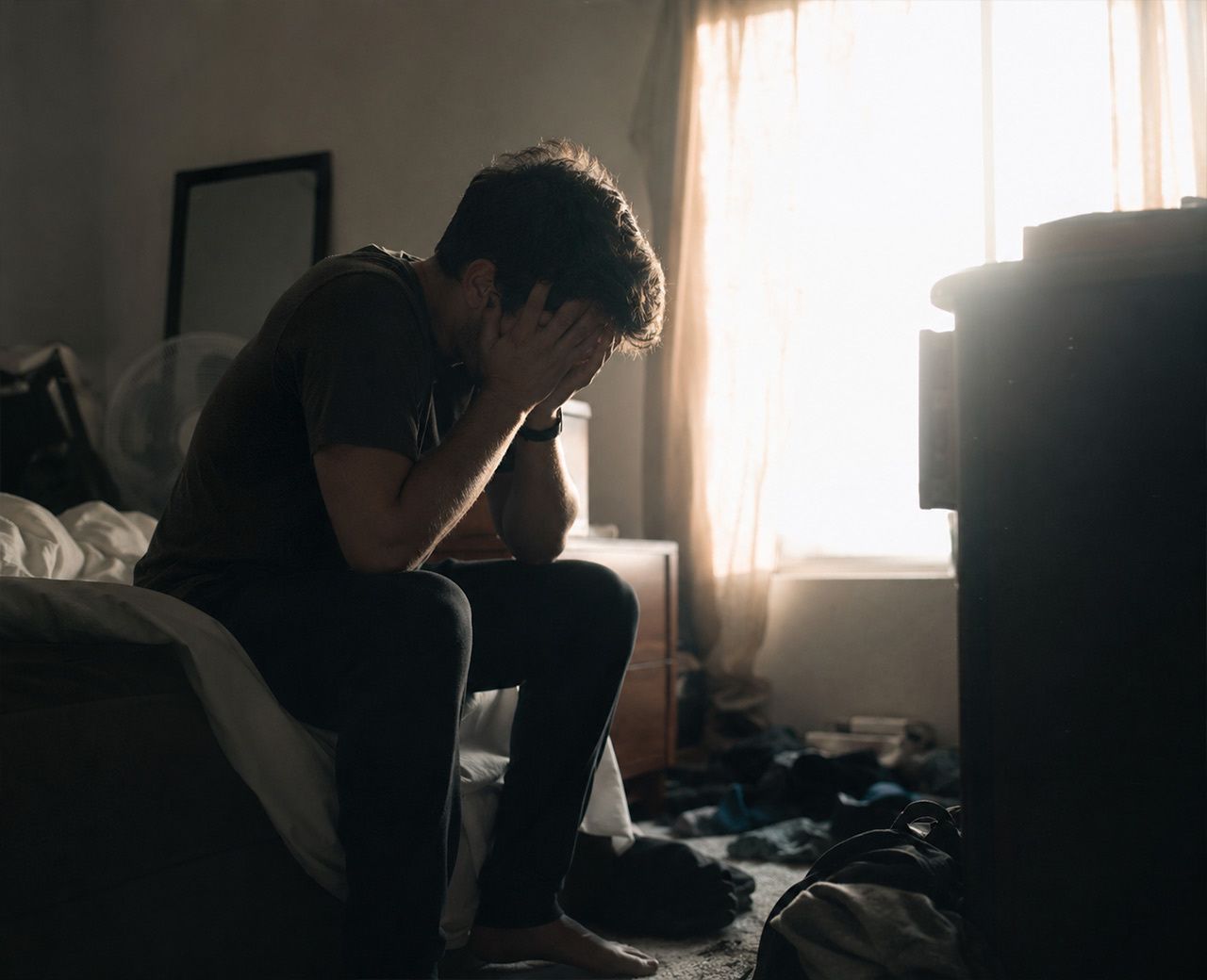 Person sitting on the edge of their bed with head in hands, harsh morning light coming through thin curtains behind them