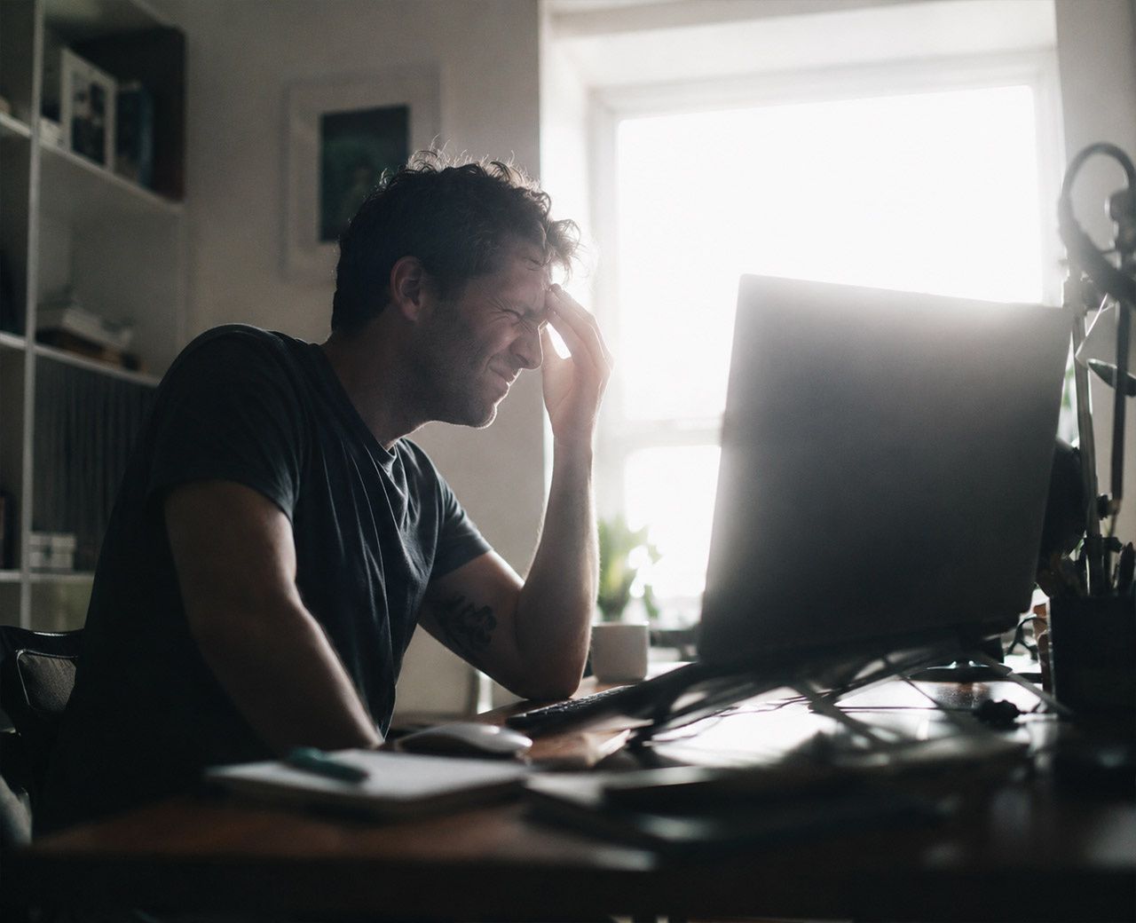 Person at a desk with a bright window behind their monitor creating harsh backlight glare, leaning forward with strained eyes