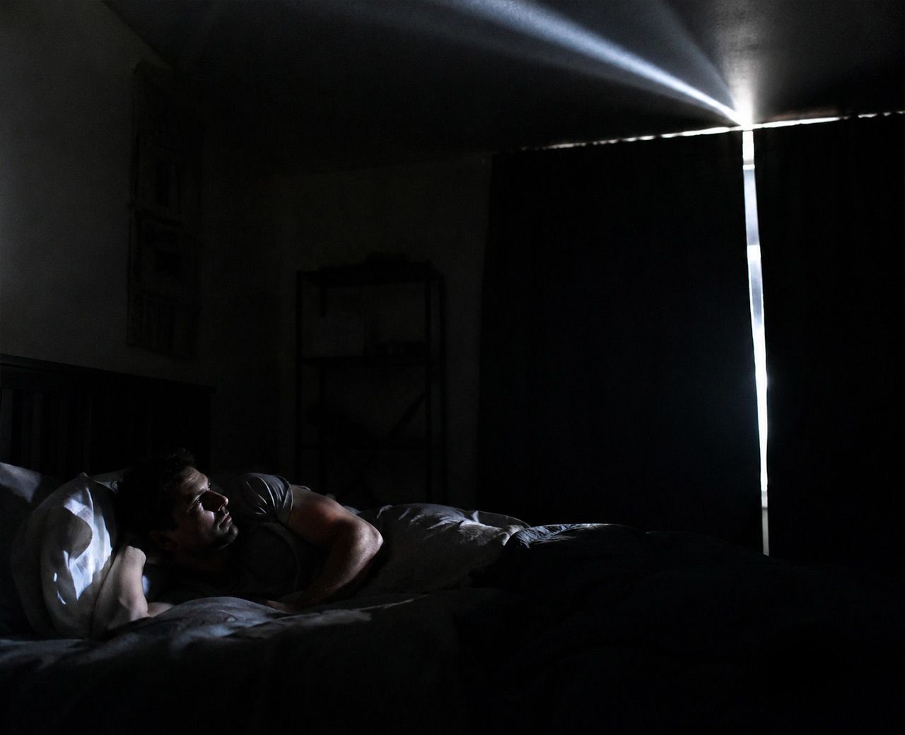 Dark bedroom at night with a bright line of streetlight cutting through a gap at the edge of drawn curtains across the bed