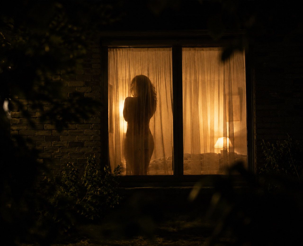 Silhouette of a person visible through light-filtering bedroom curtains from outside at night with interior lights on