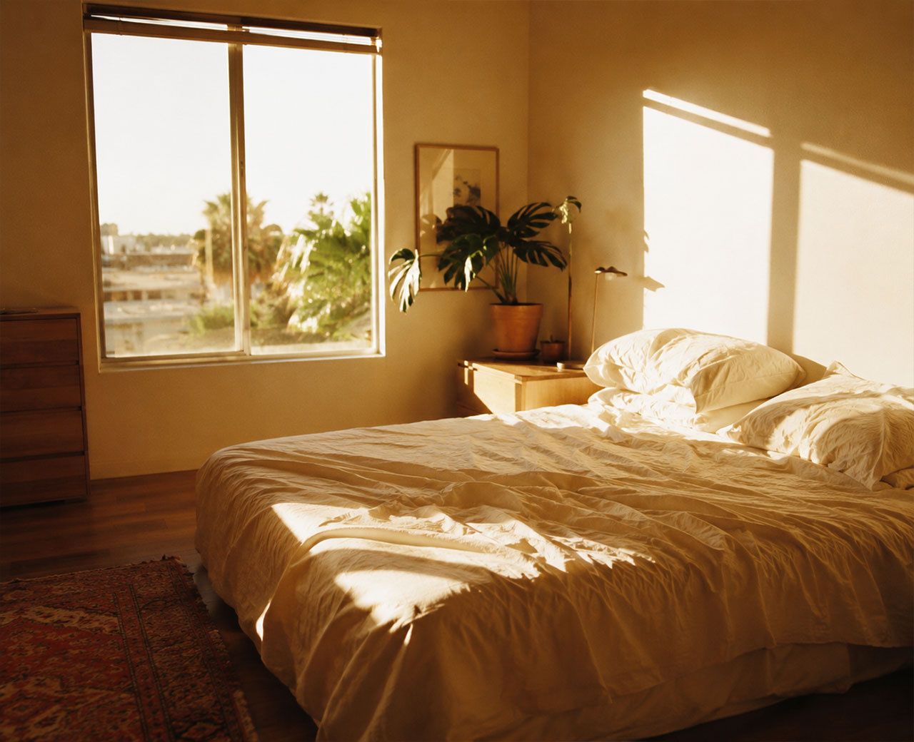 Bright bedroom interior during the day with intense direct sunlight pouring through a large uncovered window onto the bed and floor