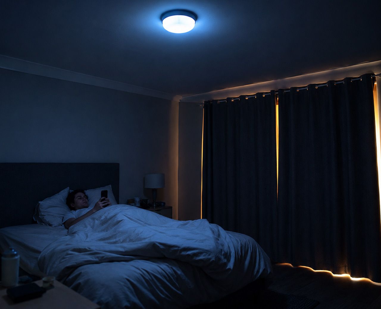  Person scrolling phone in bed with bright overhead light still on and light bleeding through curtain gaps in the background
