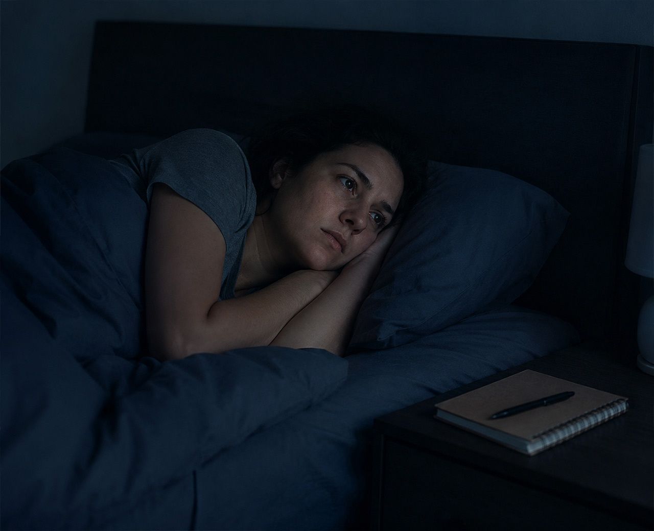 Person lying in bed in a dark room with eyes open and a tense expression, a notebook and pen on the nightstand beside them