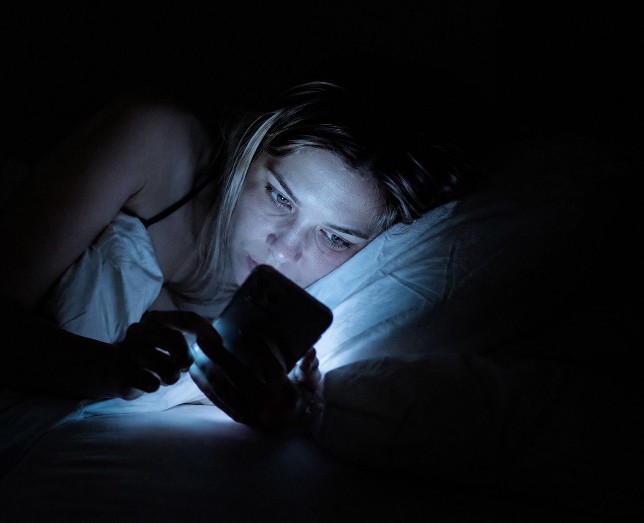 Person in bed at night scrolling their phone at full brightness, face lit by harsh blue-white screen light in a pitch black room
