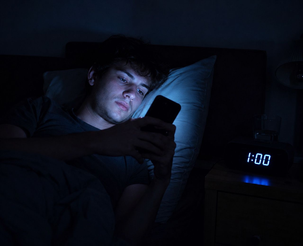 Person checking their phone in bed late at night with the clock showing 1am in an otherwise dark bedroom