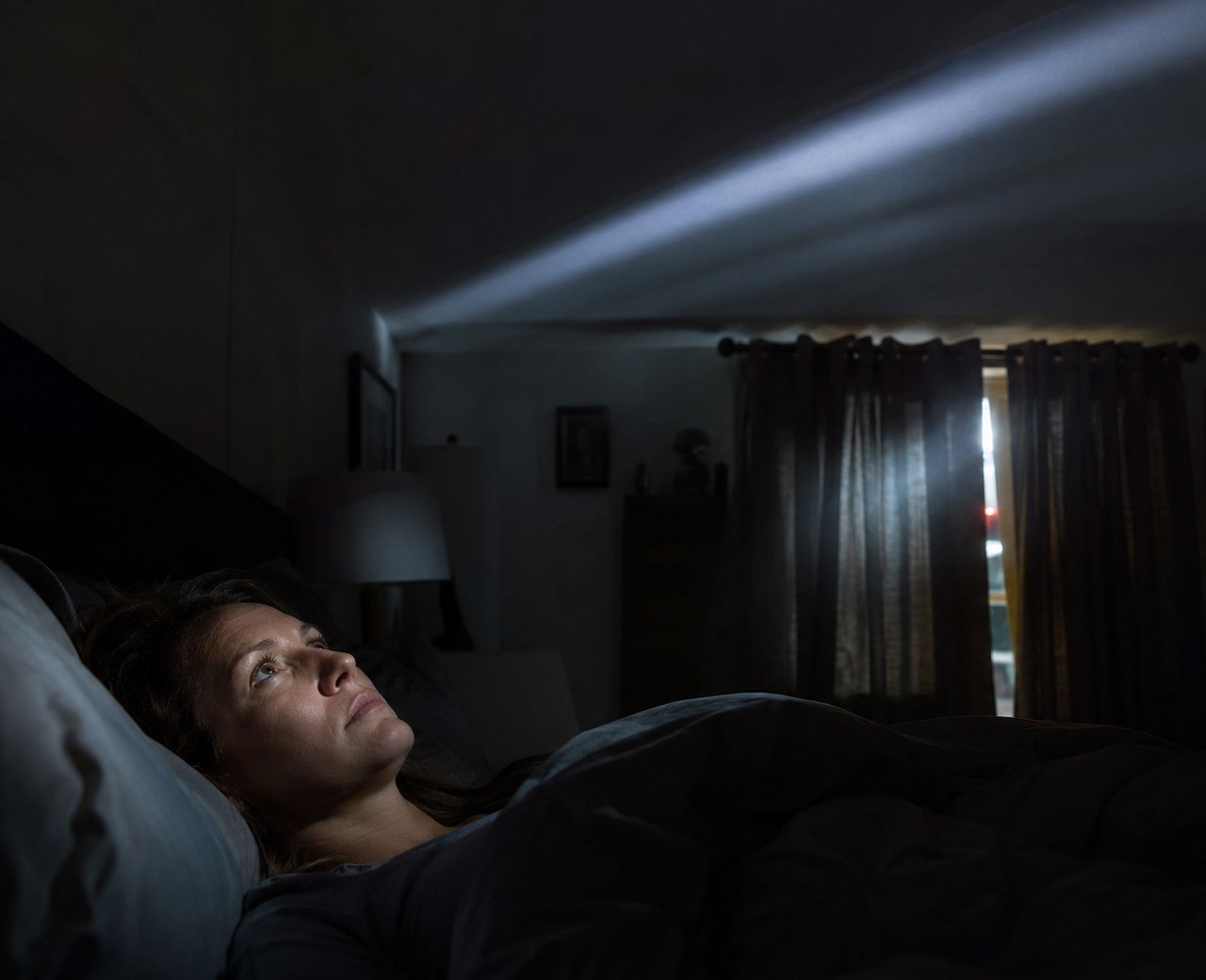 Person lying awake in bed as headlights sweep across the ceiling through a gap in the curtains suggesting street noise disruptions