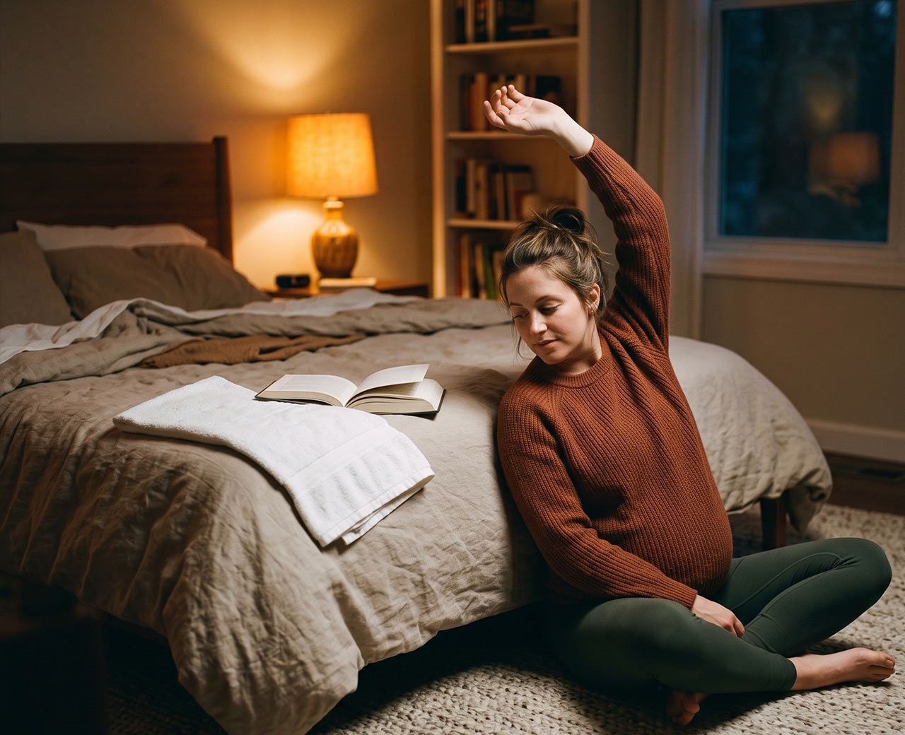 Pregnant woman doing gentle stretches beside her bed in a dimly lit room before sleep