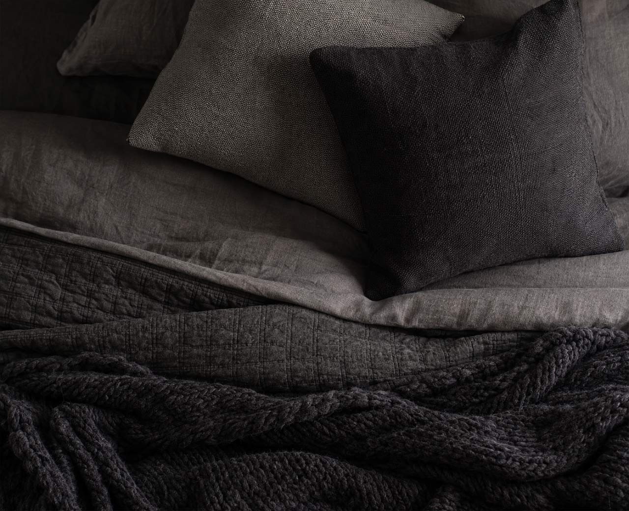 Layered dark bedding showing linen, knit and quilted textures in gray and black tones