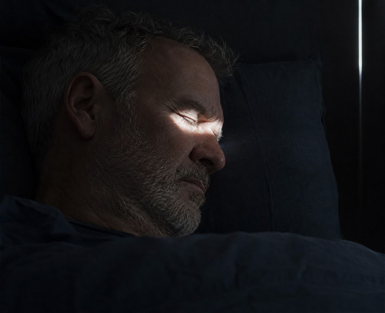 Narrow beam of dawn light falling across the face of a sleeping older man in a dark bedroom