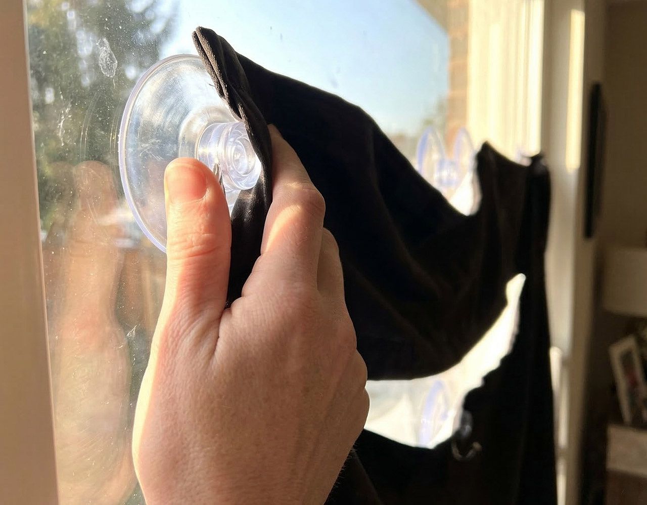 Close-up of a hand pressing a suction cup against a window pane to attach a portable blackout curtain with daylight visible through the glass